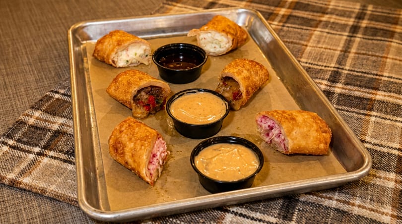 Six crispy egg rolls with various fillings and three dipping sauces on a metal tray.