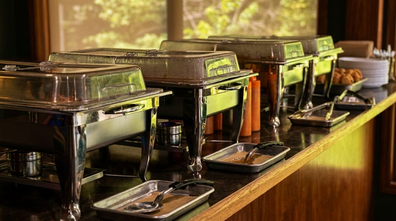 A buffet line with several silver chafing dishes, serving utensils, plates, and pastries.