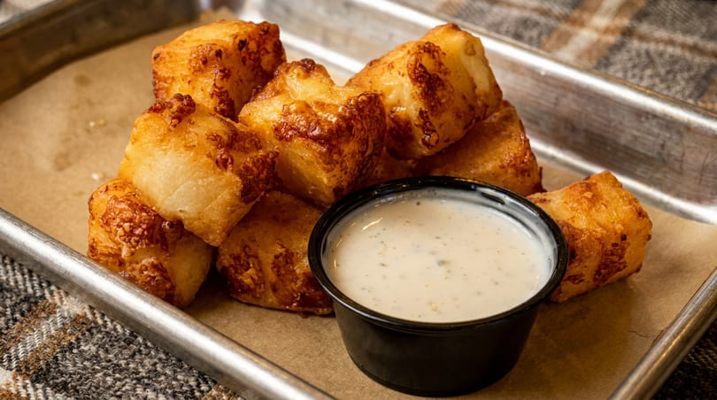 Golden-brown fried cheese curds with a side of ranch dressing on a metal tray.