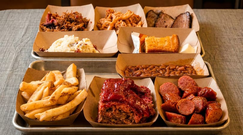 A metal tray holds nine paper boats filled with various barbecue dishes and sides.