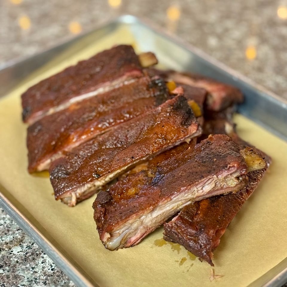 Close-up of several glistening, saucy BBQ ribs on a metal tray lined with parchment paper.